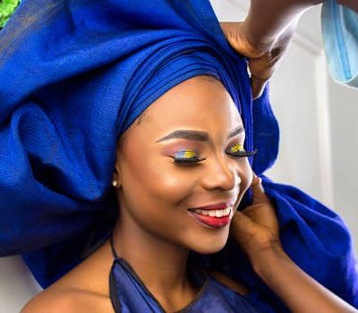Smiling Nigerian woman in blue headwrap getting ready with vivid makeup.