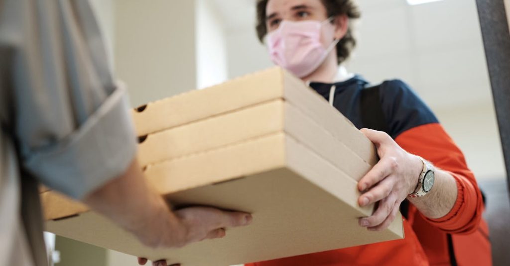 Delivery worker handing over pizzas while wearing a protective mask indoors, illustrating safe delivery practices during a pandemic.